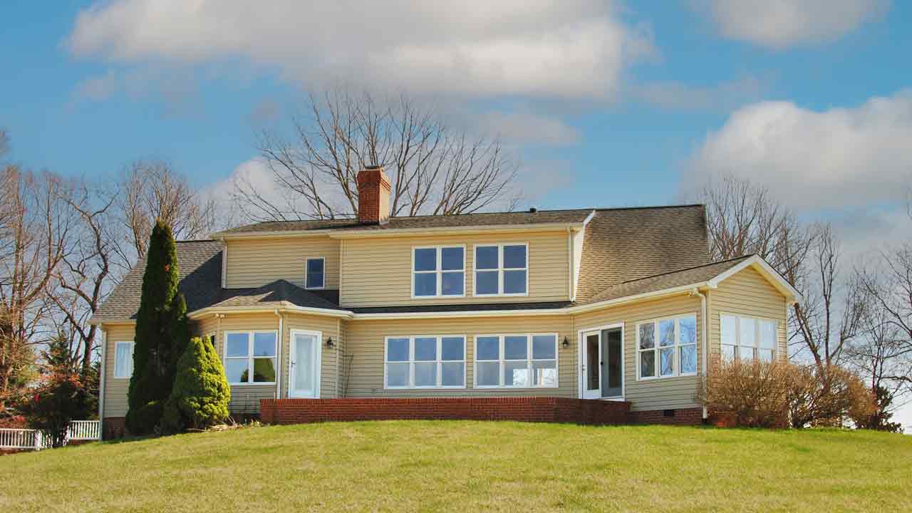 A charming two-story country home with beige siding, a brick foundation, and large windows that flood the interior with natural light. The house features a central brick chimney, a sunroom extension, and a welcoming entrance. Set on a gently sloping lawn with neatly trimmed shrubs and evergreens, the property offers a peaceful rural setting under a bright blue sky with scattered clouds.