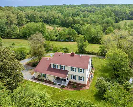Aerial view of renovated farmhouse with red roof and covered porch at 1701 Harrington Road in Gordonsville, VA, surrounded by mature trees and open pasture.