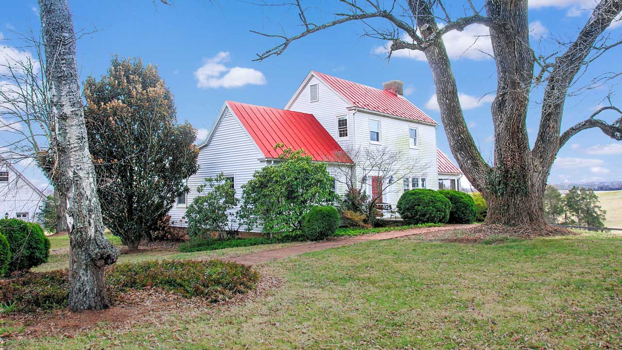 A picturesque historic farmhouse with white siding and a bold red metal roof, set amidst lush greenery. This timeless home showcases classic architectural details, including a central chimney, multiple gables, and a charming red front door. A brick pathway winds through neatly manicured bushes and mature trees, leading to the inviting entrance. A towering, weathered tree in the foreground adds character, while the bright blue sky with scattered clouds enhances the idyllic country setting.
