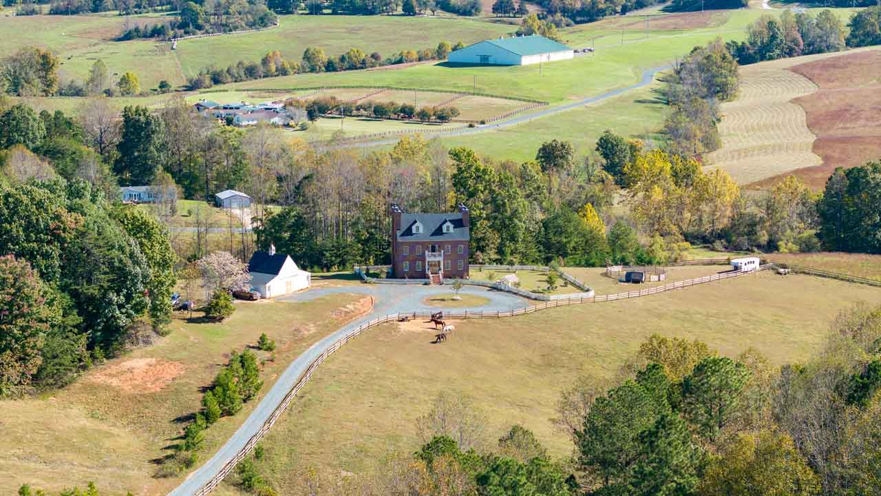 Aerial view of an elegant brick estate with a grand symmetrical facade, surrounded by rolling pastures and equestrian facilities. A long, winding gravel driveway leads to the circular entrance with a landscaped fountain. The fenced property includes a riding area with horses grazing, a guest house, and a horse trailer. The scenic countryside setting features open fields, a large barn in the distance, and wooded areas enhancing the rural charm.