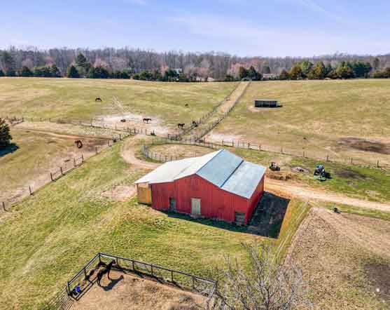 Aerial view of a horse farm with a red barn, fenced paddocks, grazing horses, and rolling pastures.