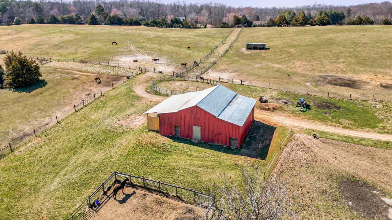 Aerial view of a red barn on a 71-acre farm in Louisa County, VA, surrounded by fenced pastures with grazing horses, open fields, and farm equipment visible near the barn.