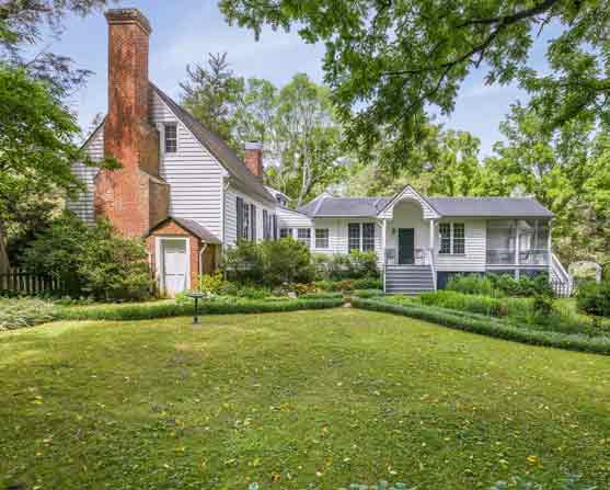 Historic white clapboard home with prominent brick chimneys and a gabled addition, surrounded by lush gardens and mature trees, featuring a manicured lawn with formal boxwood edging.