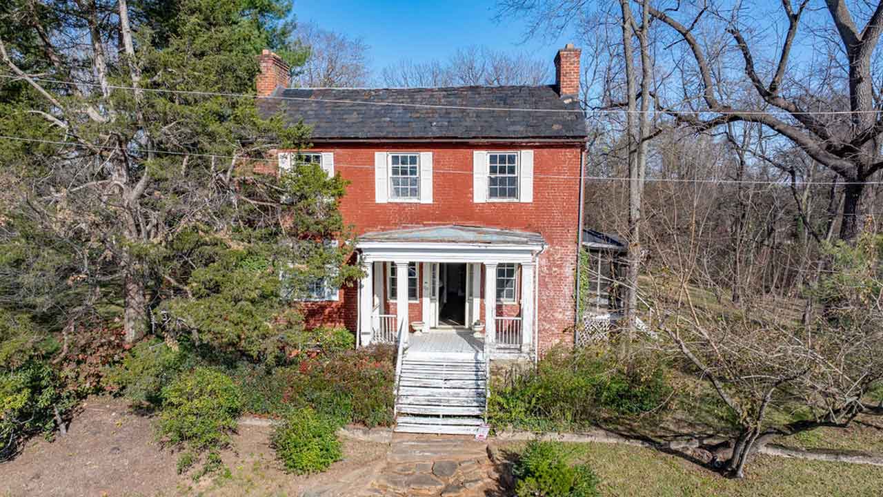 A historic two-story red brick home with white shutters and a charmingly aged front porch, complete with wooden stairs leading to the entrance. Featuring a symmetrical facade, a slate roof, and twin brick chimneys, the home exudes classic appeal. Mature trees and lush overgrown shrubs frame the property, enhancing its character and offering a glimpse of its restoration potential. The open front door invites curiosity, hinting at the history within.