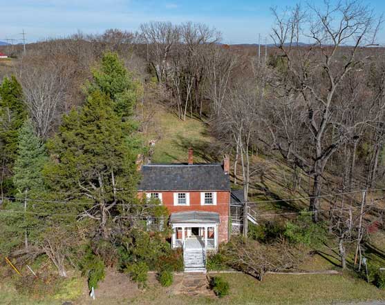 An aerial view of a historic two-story brick house with a black roof, nestled among mature trees and surrounded by a natural landscape. The front of the house features a small covered porch with white columns and a staircase leading to the entrance. The surrounding area includes bare trees, open fields, and a backdrop of rolling hills under a clear sky, giving the property a rustic and timeless charm.