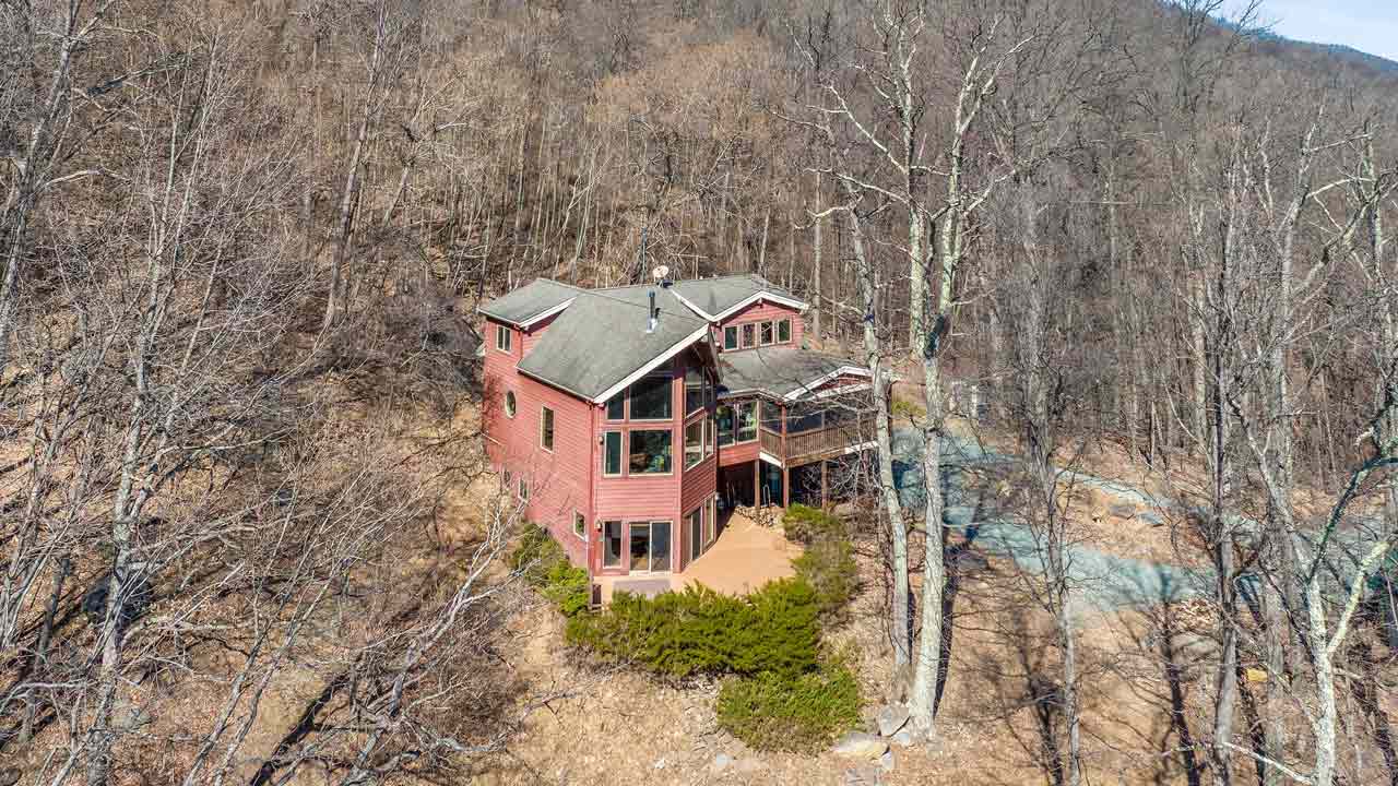 A historic two-story red brick home with white shutters and a charmingly aged front porch, complete with wooden stairs leading to the entrance. Featuring a symmetrical facade, a slate roof, and twin brick chimneys, the home exudes classic appeal. Mature trees and lush overgrown shrubs frame the property, enhancing its character and offering a glimpse of its restoration potential. The open front door invites curiosity, hinting at the history within.