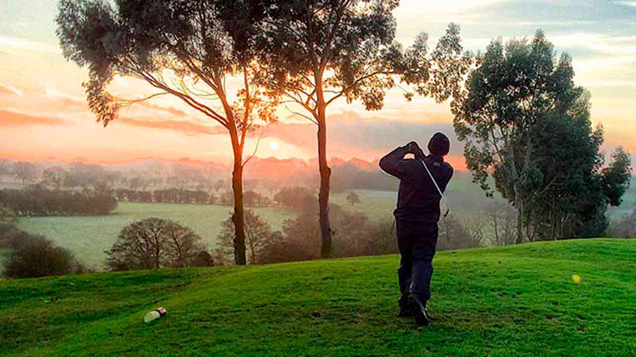 A golfer takes a swing at sunrise on a lush, green fairway surrounded by tall trees and misty countryside, capturing the peaceful beauty of early morning golf in Central Virginia.