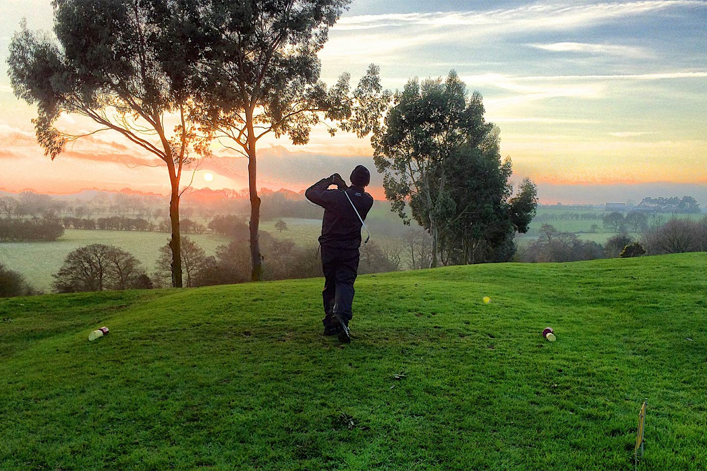 Golfer in mid-swing on a lush green course, surrounded by trees and an expansive countryside view, with a vibrant sunset casting warm hues over the horizon.