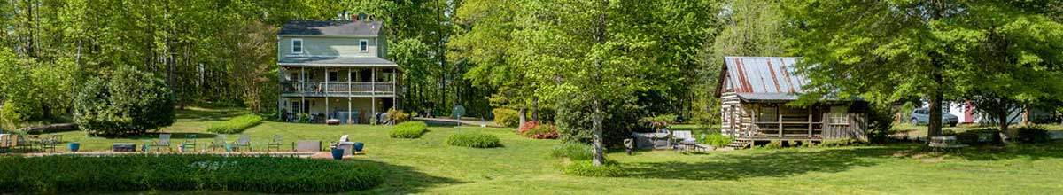 Charlottesville country property featuring a charming farmhouse with a wraparound porch, historic log cabin, outdoor living areas, and wooded landscape.